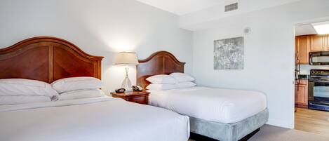 A hotel room with two beds, a nightstand with a lamp, a ceiling fan, and a view of the kitchen.
