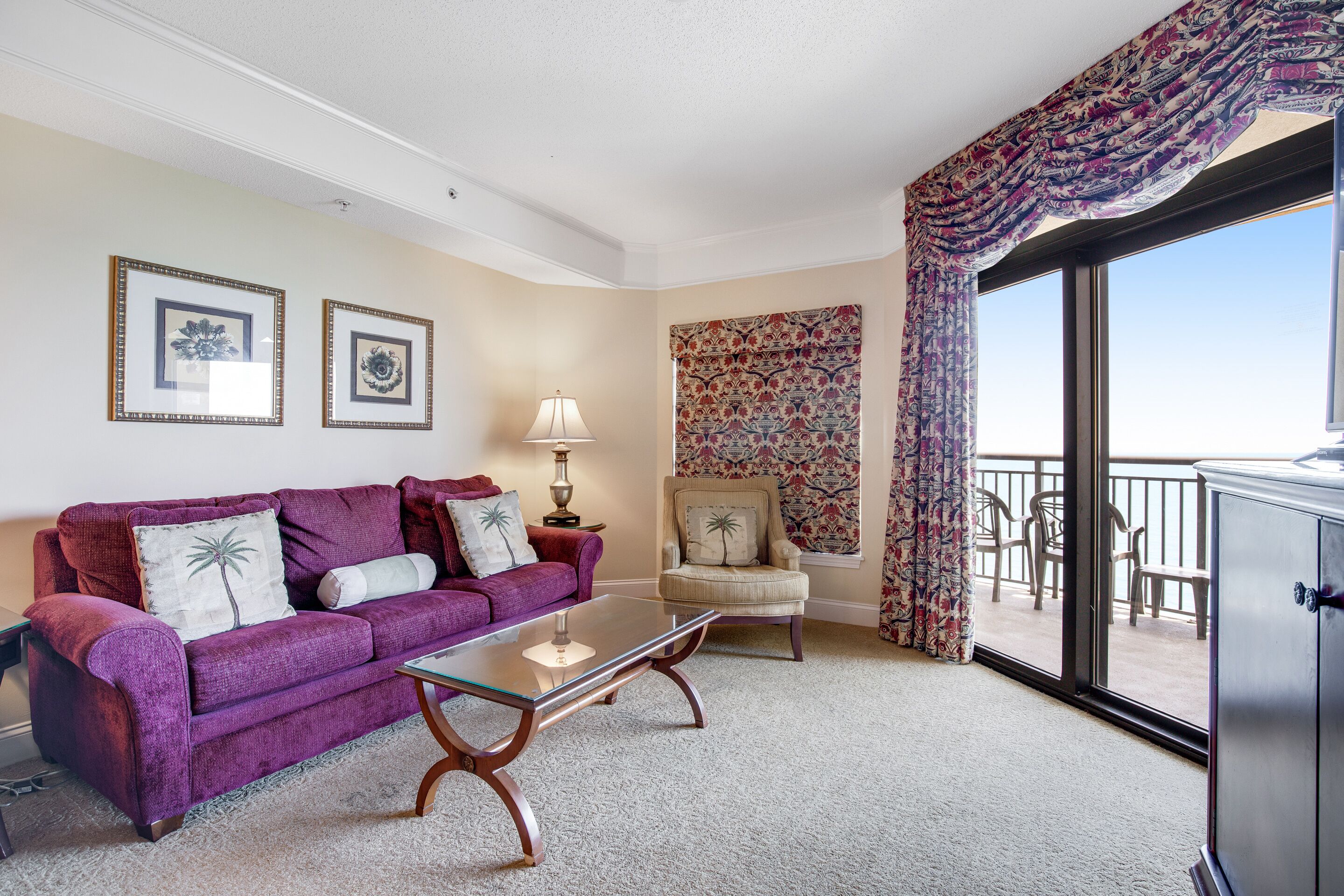 Uma sala de estar com um sofá roxo, uma mesa de centro de vidro, uma poltrona bege e uma cortina estampada.