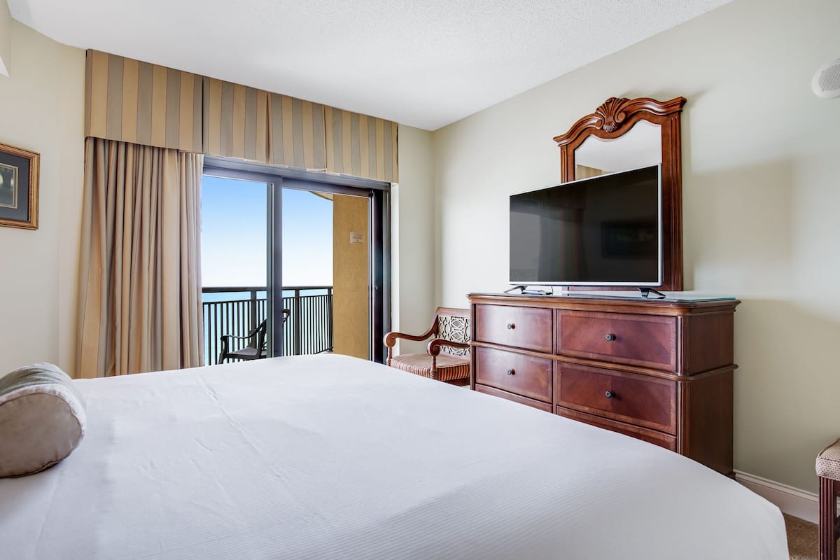 Une chambre d’hôtel avec un grand lit, une commode en bois, une télévision et un balcon avec une chaise.