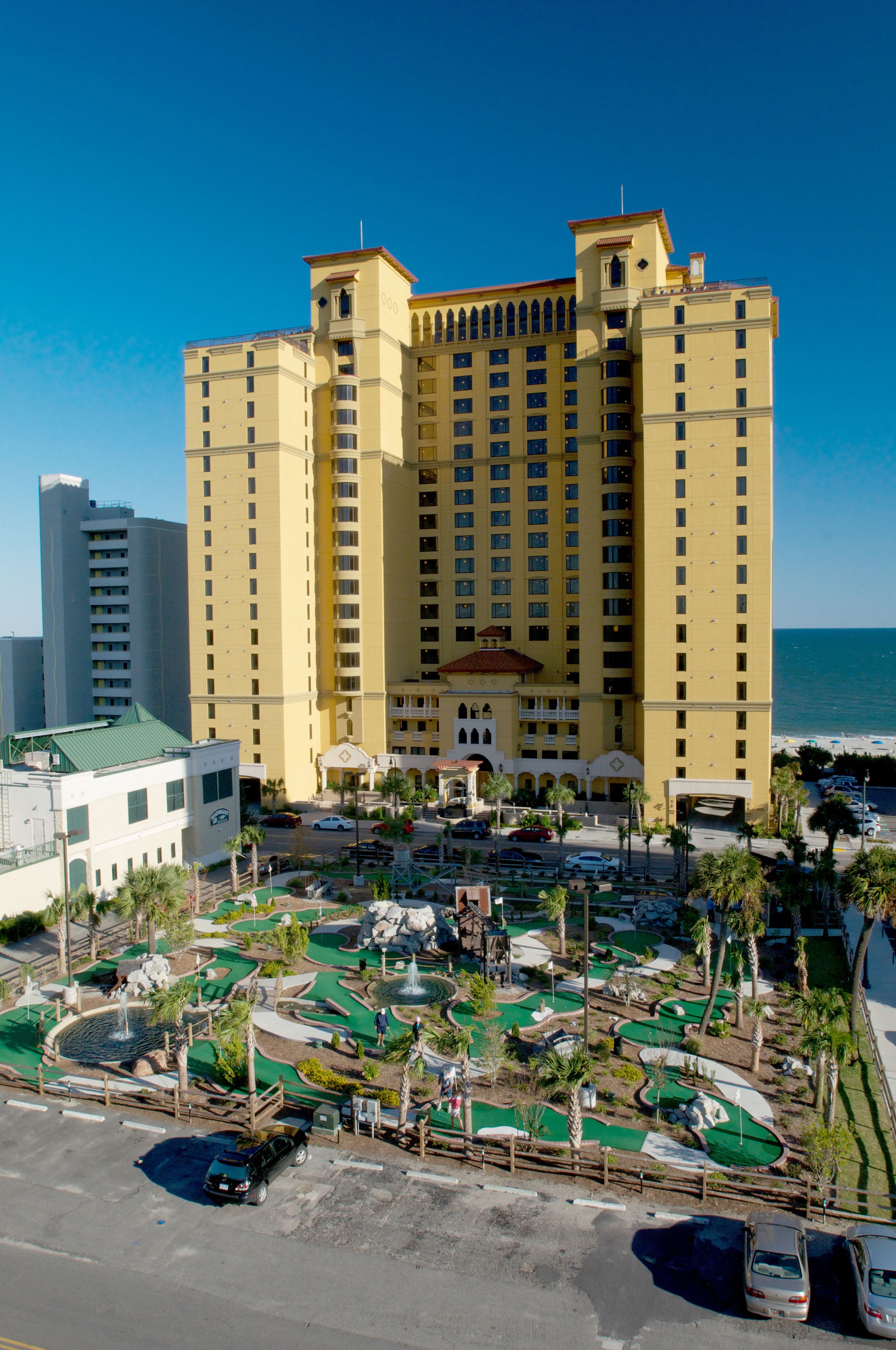 A large hotel with multiple floors, a parking area, and a pool complex.