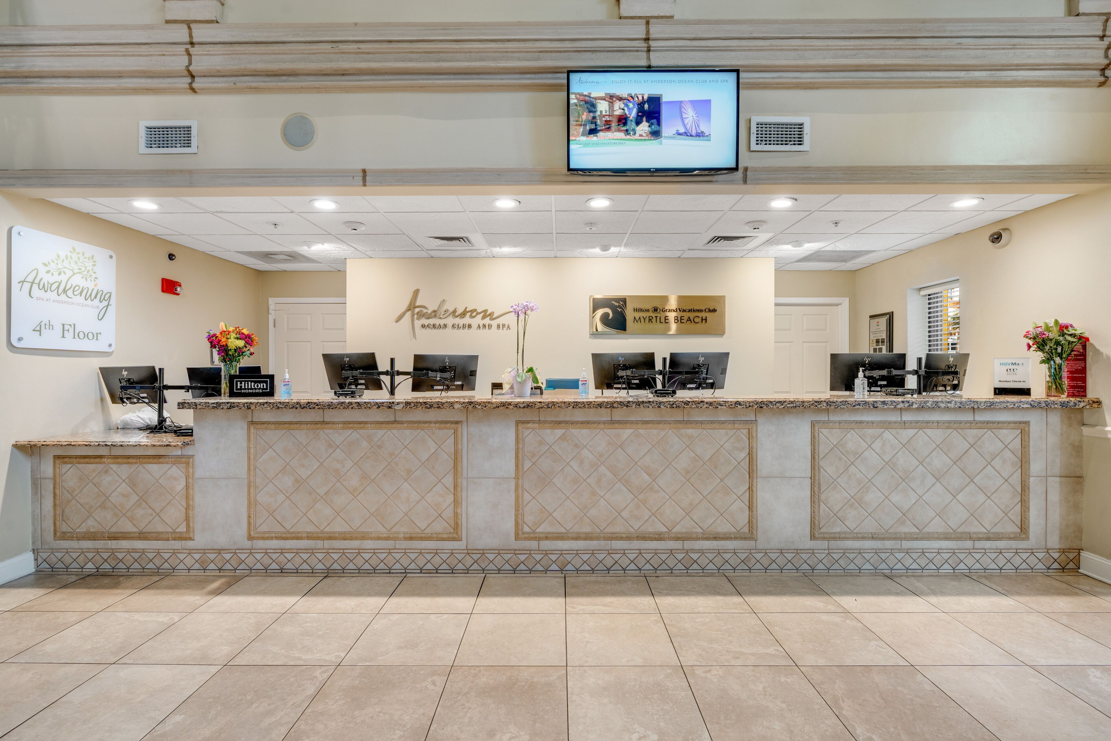 A hotel reception area with a tiled floor, a reception desk with a television mounted above it, and signs indicating the hotel's name and location.
