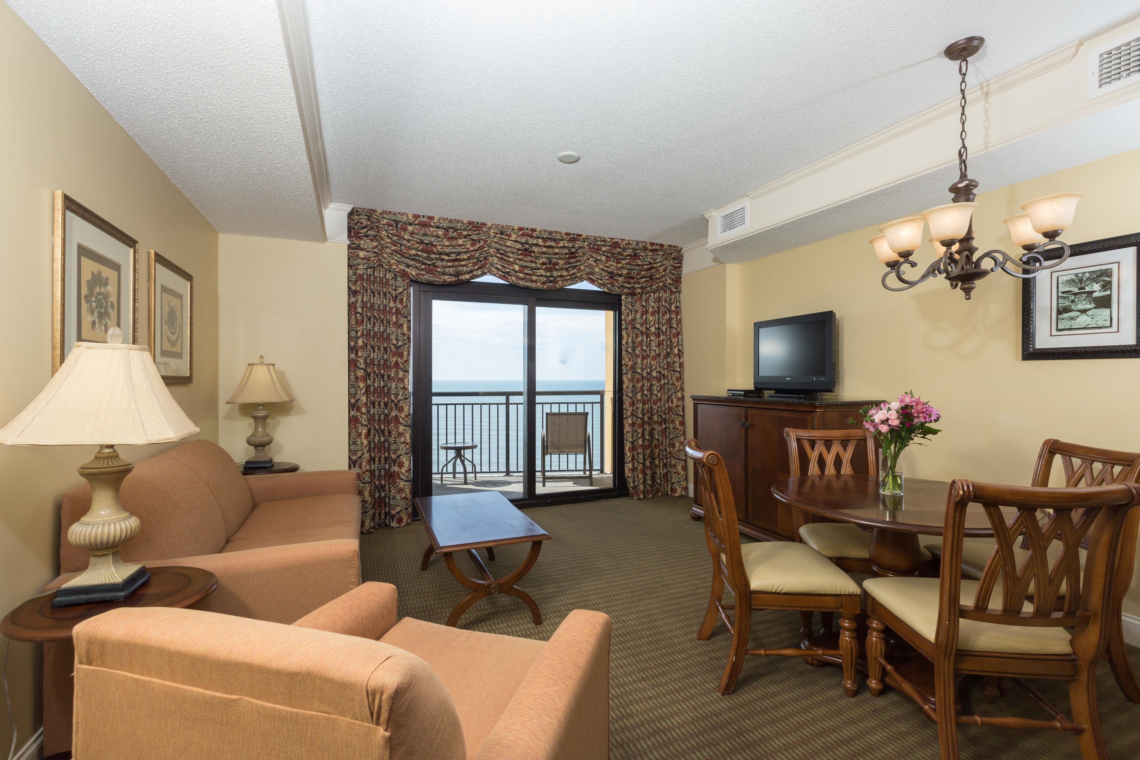 A hotel room with a sofa, armchair, dining table, chairs, a TV, and a balcony with a view.