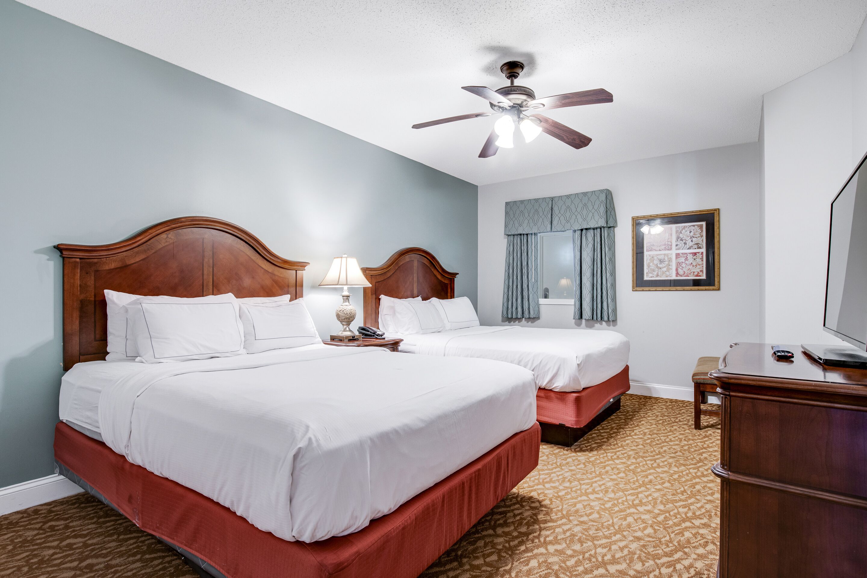 A hotel room with two beds, a ceiling fan, a desk, and a television.