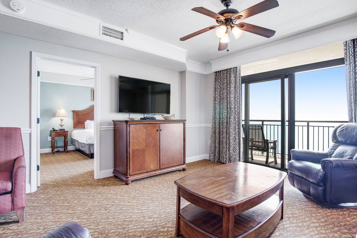 Une chambre d’hôtel comprenant un lit, une télévision, une armoire en bois, un fauteuil en cuir, une table basse en bois et un balcon avec vue.