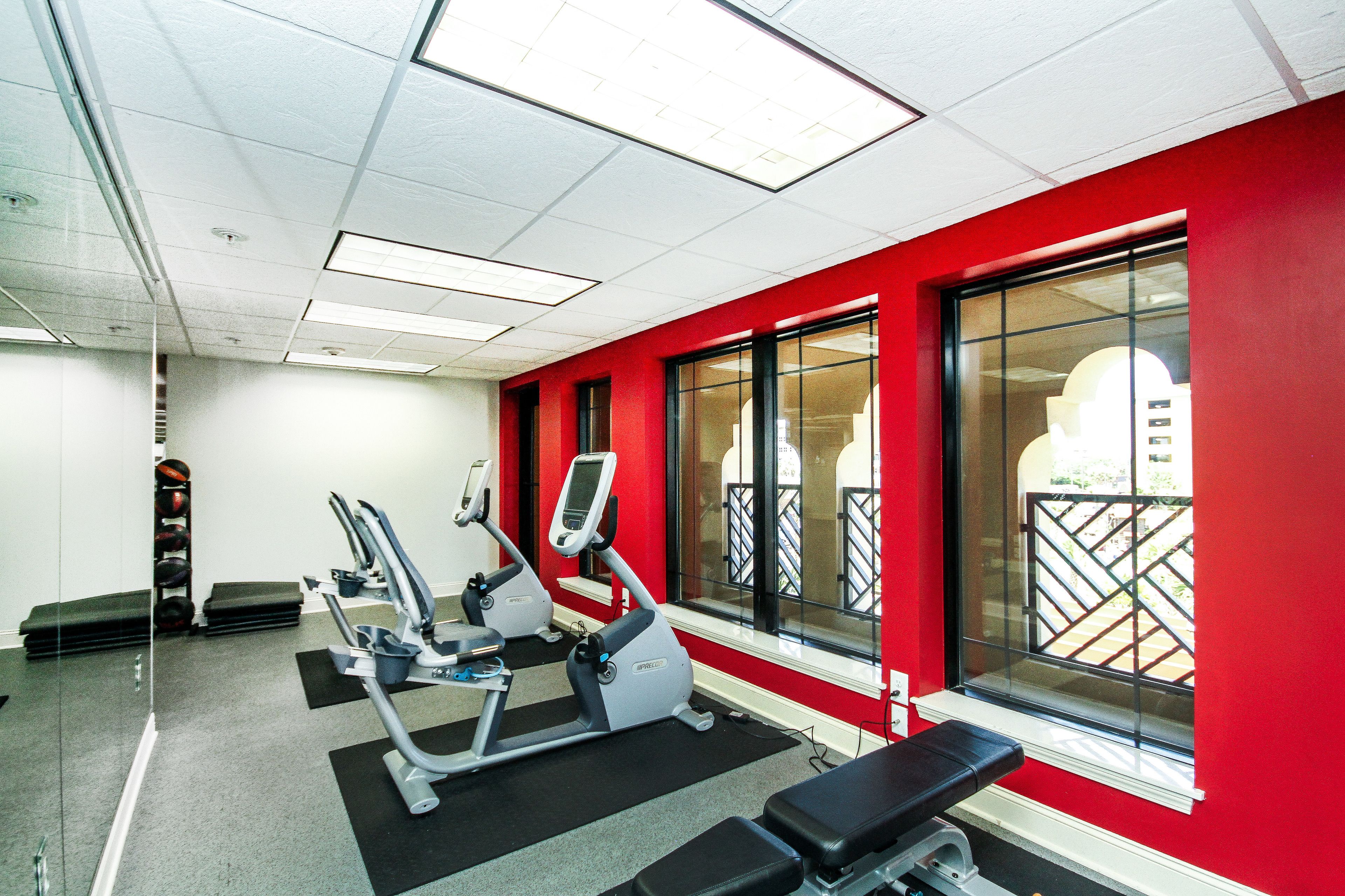 Un gimnasio con equipo de ejercicio, ventanas amplias y una pared roja.