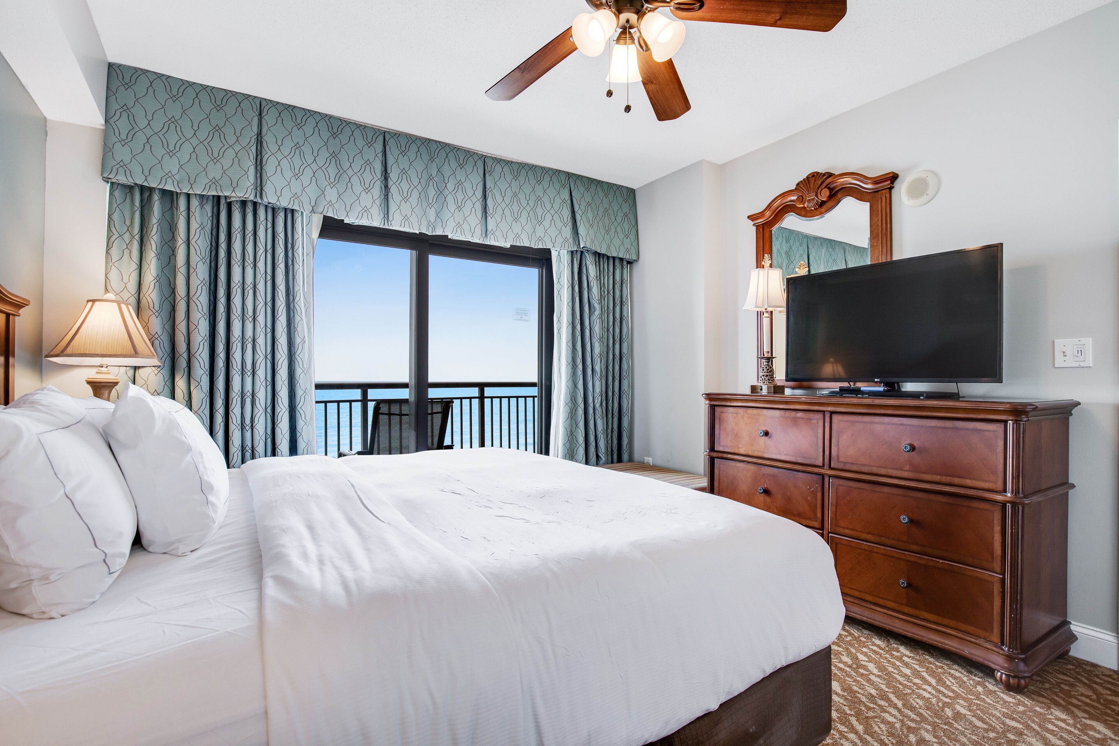 Een hotelkamer met een groot bed, een houten dressoir, een televisie en een balkon met gordijnen.