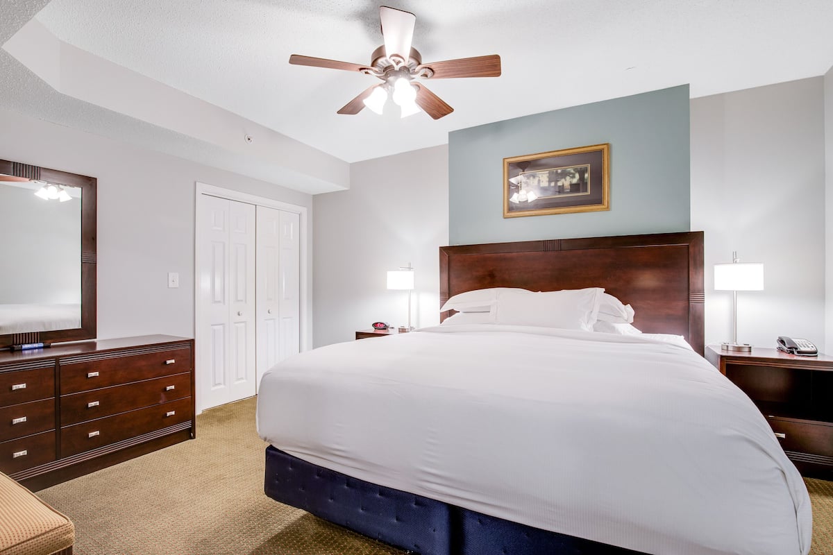 Une chambre d’hôtel avec un grand lit, une tête de lit en bois, un ventilateur de plafond, une table de chevet avec une lampe et une commode.