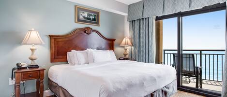 A large bed with white linens, a wooden headboard, two bedside tables with lamps, a framed picture on the wall, and a balcony with a view of the ocean.