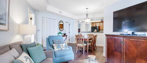 Una sala de estar con sofá, sillón y mesa de centro, un comedor con mesa y sillas, y una cocina al fondo.