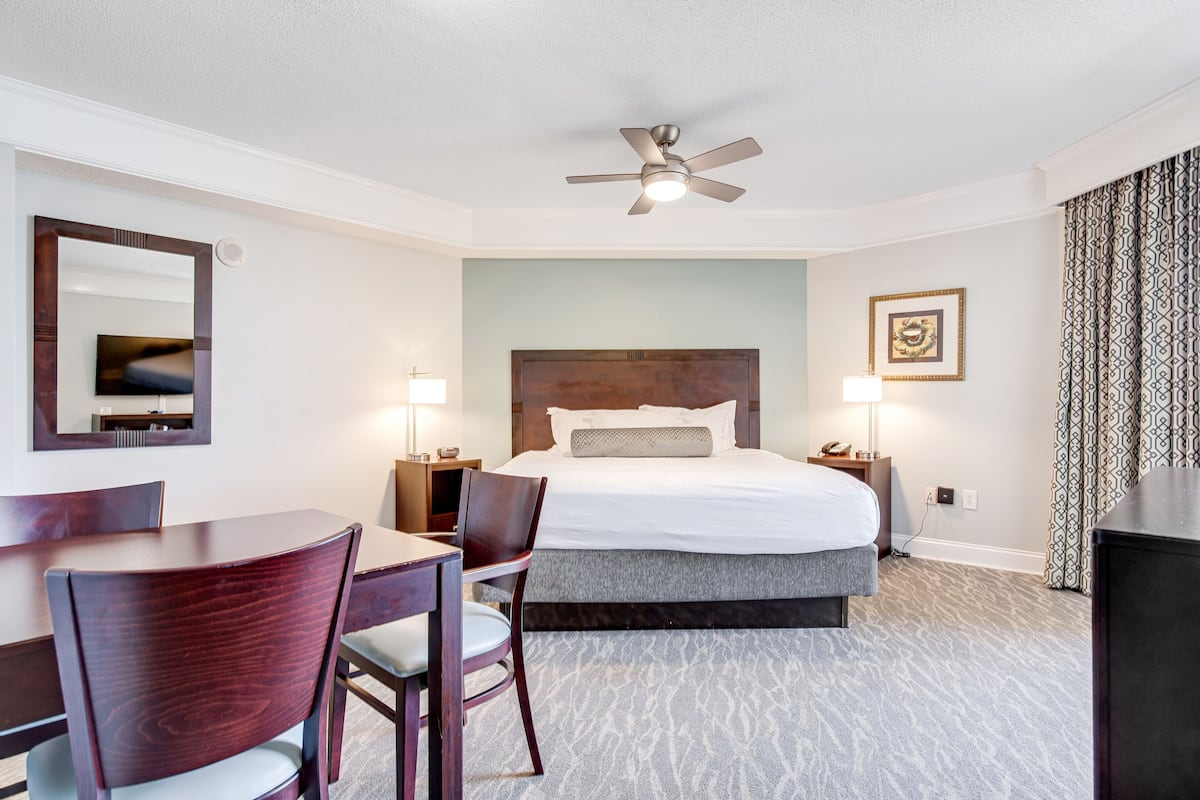 Une chambre d’hôtel avec un lit, un bureau, une chaise, un ventilateur de plafond, un miroir et un tableau encadré au mur.
