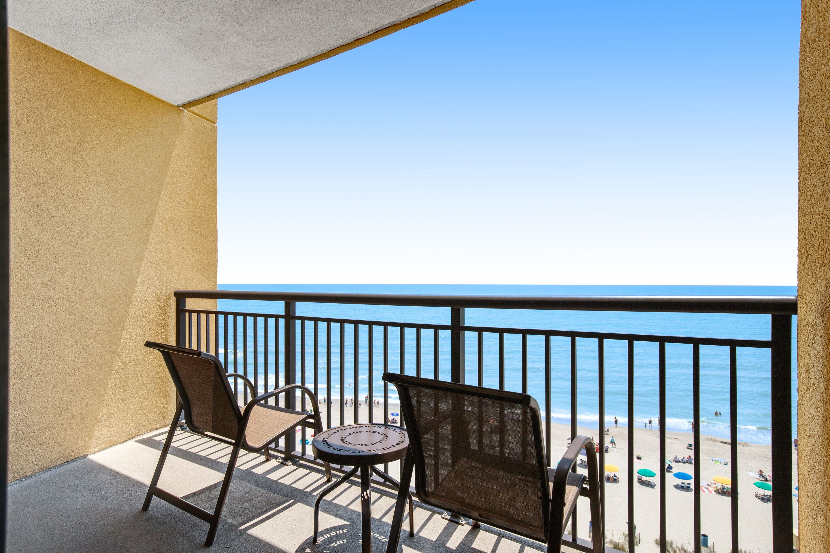 A balcony with two chairs and a small table, overlooking a beach with umbrellas and people.