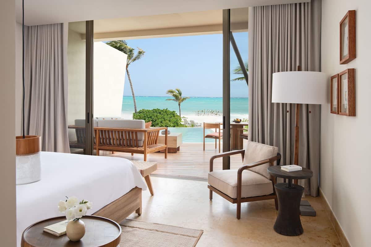 A hotel room with a bed, a chair, a small table, and a view of the beach and ocean.