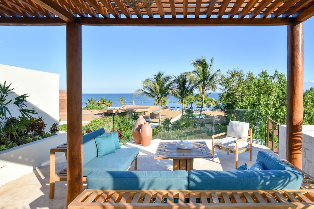 A wooden pergola with a view of the ocean, a sofa, a chair, and a table with a cushion.