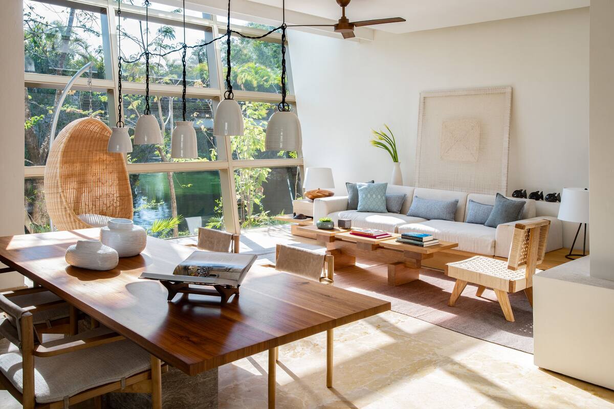 A spacious living room with a large window, a wooden dining table, a sofa, and a hanging chair.