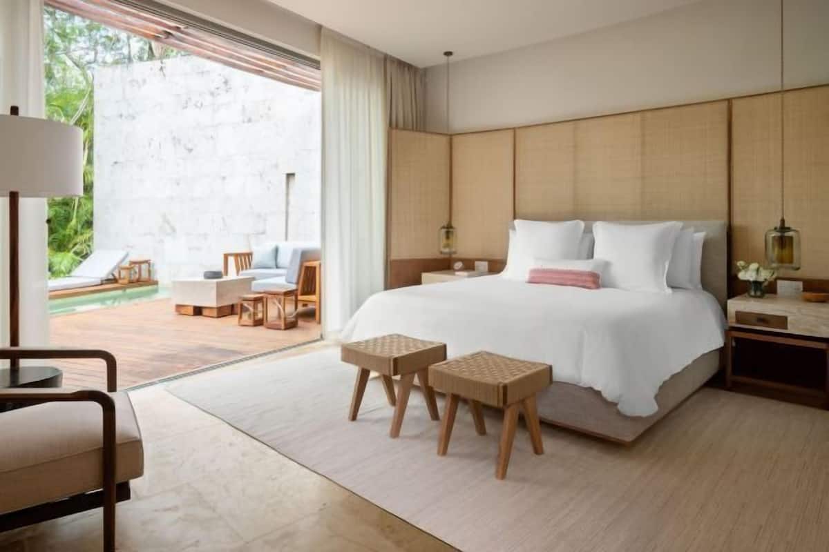 A modern hotel room with a large bed, a sitting area with a table and chairs, and a view of a garden through a sliding door.