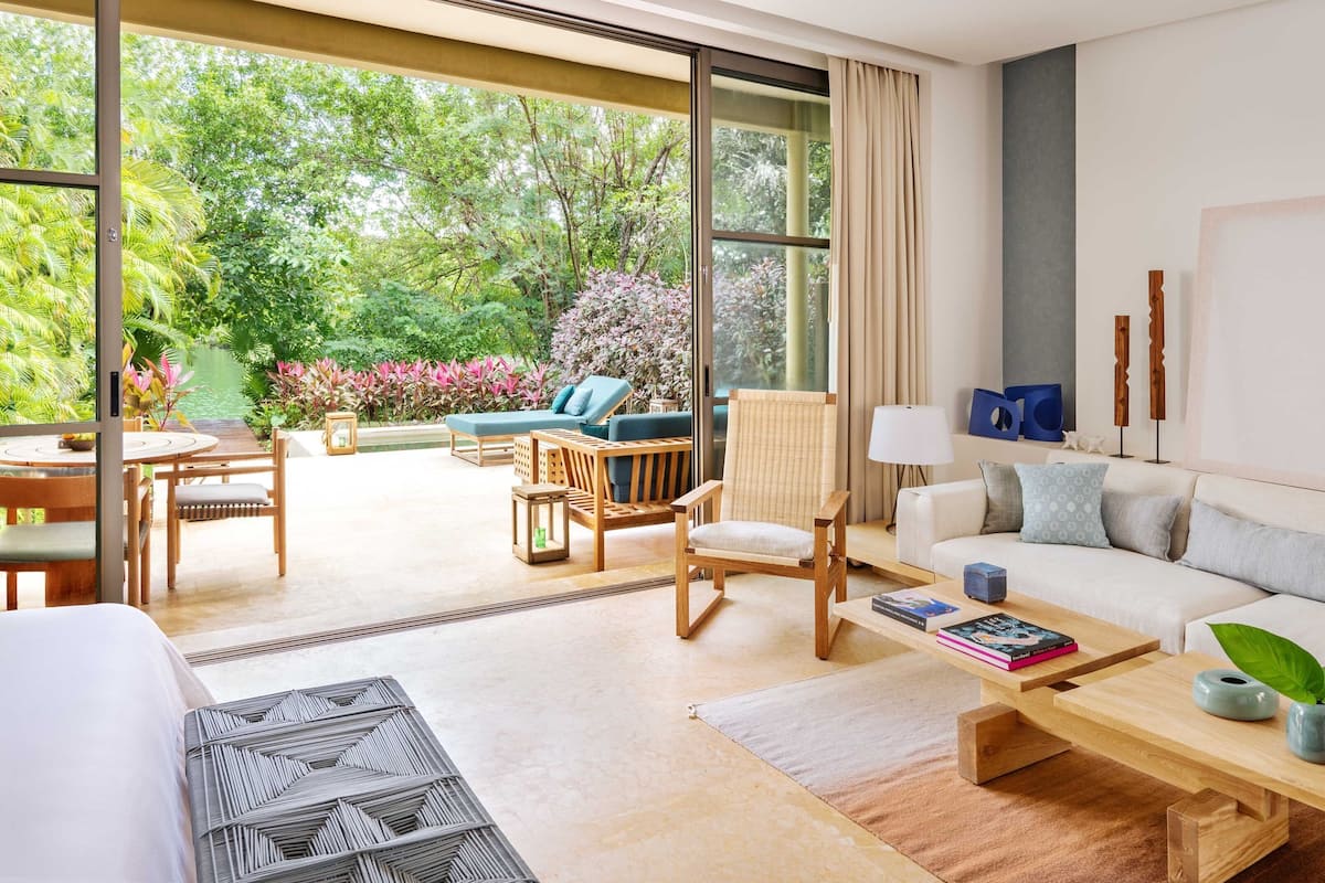 A spacious living room with a large sliding glass door leading to a lush garden, a comfortable sofa, a wooden coffee table, and a dining area with a table and chairs.