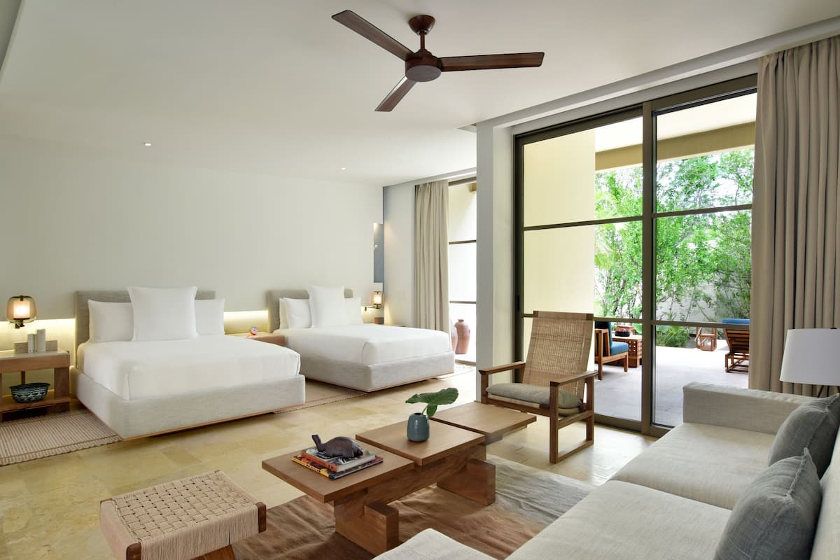 A spacious hotel room with a large bed, a sofa, a chair, and a coffee table. There is a ceiling fan, a window with curtains, and a view of greenery outside.