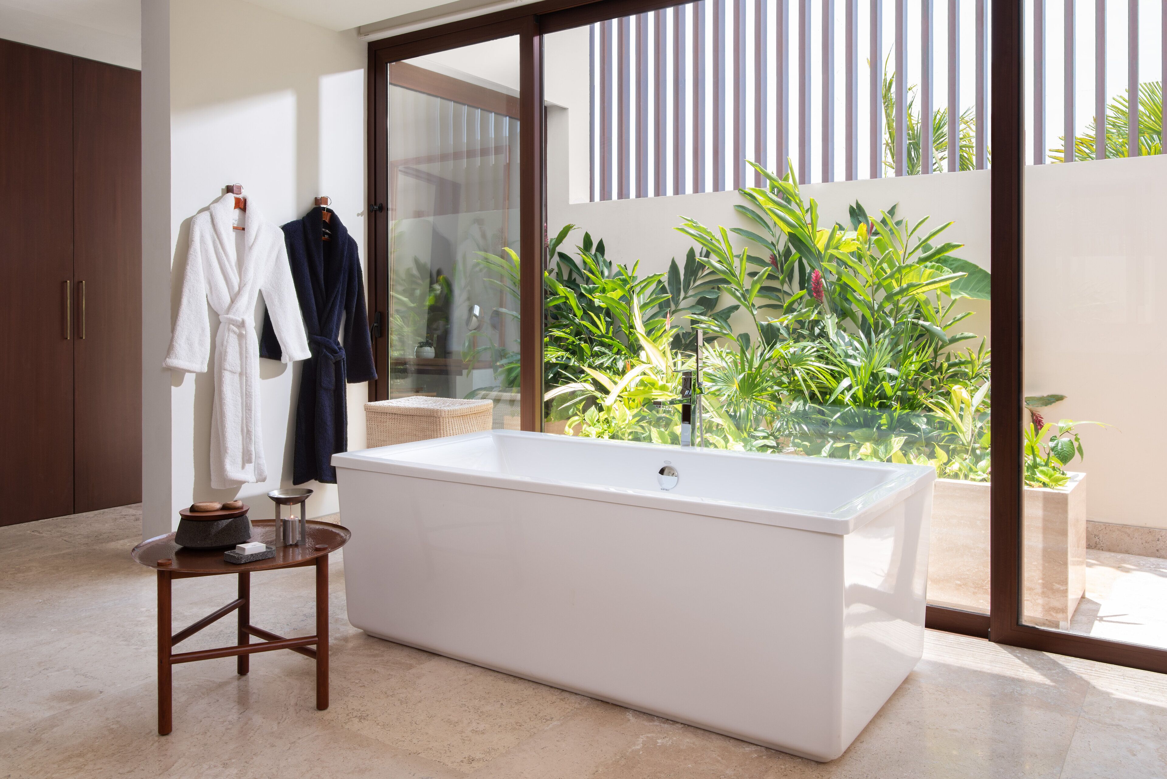 Un baño moderno con una bañera grande, un taburete de madera y una puerta de cristal que da a un balcón con plantas.