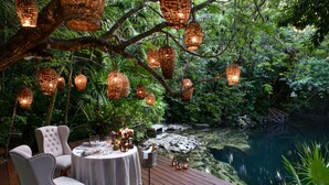 An outdoor dining area with wicker lanterns, a round table, and chairs, overlooking a pond surrounded by lush greenery. - Rosewood Mayakoba (Playa del Carmen)