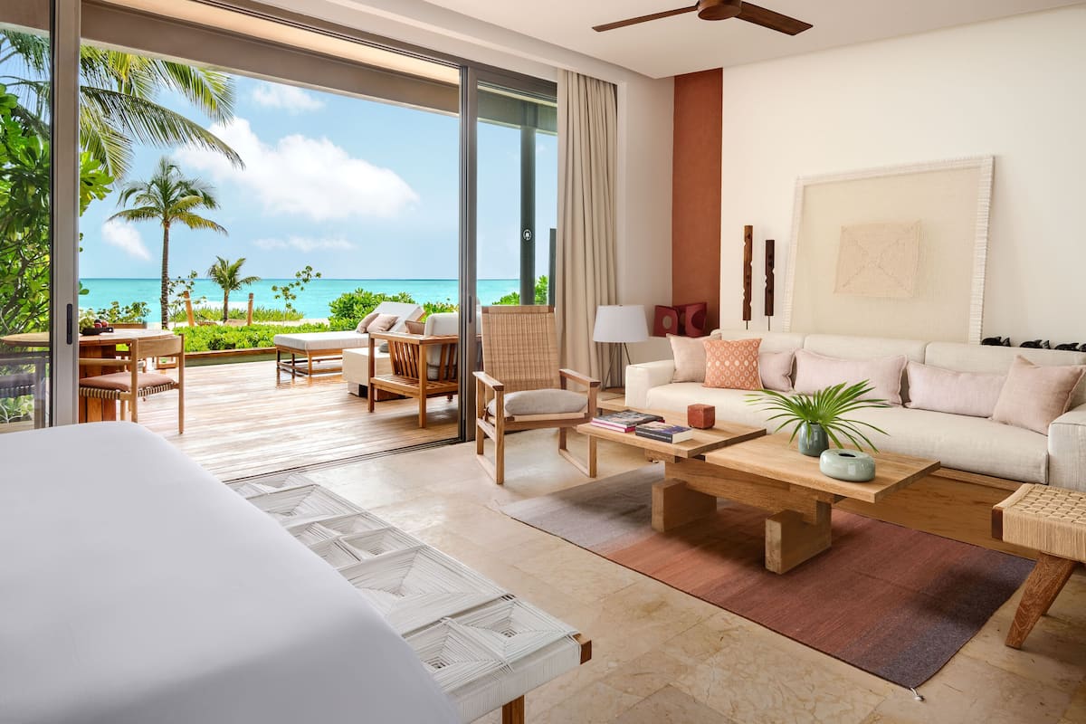 A modern hotel room with a large sliding glass door leading to a balcony with a view of the ocean. Inside, there is a bed, a sofa, a coffee table, and a chair.