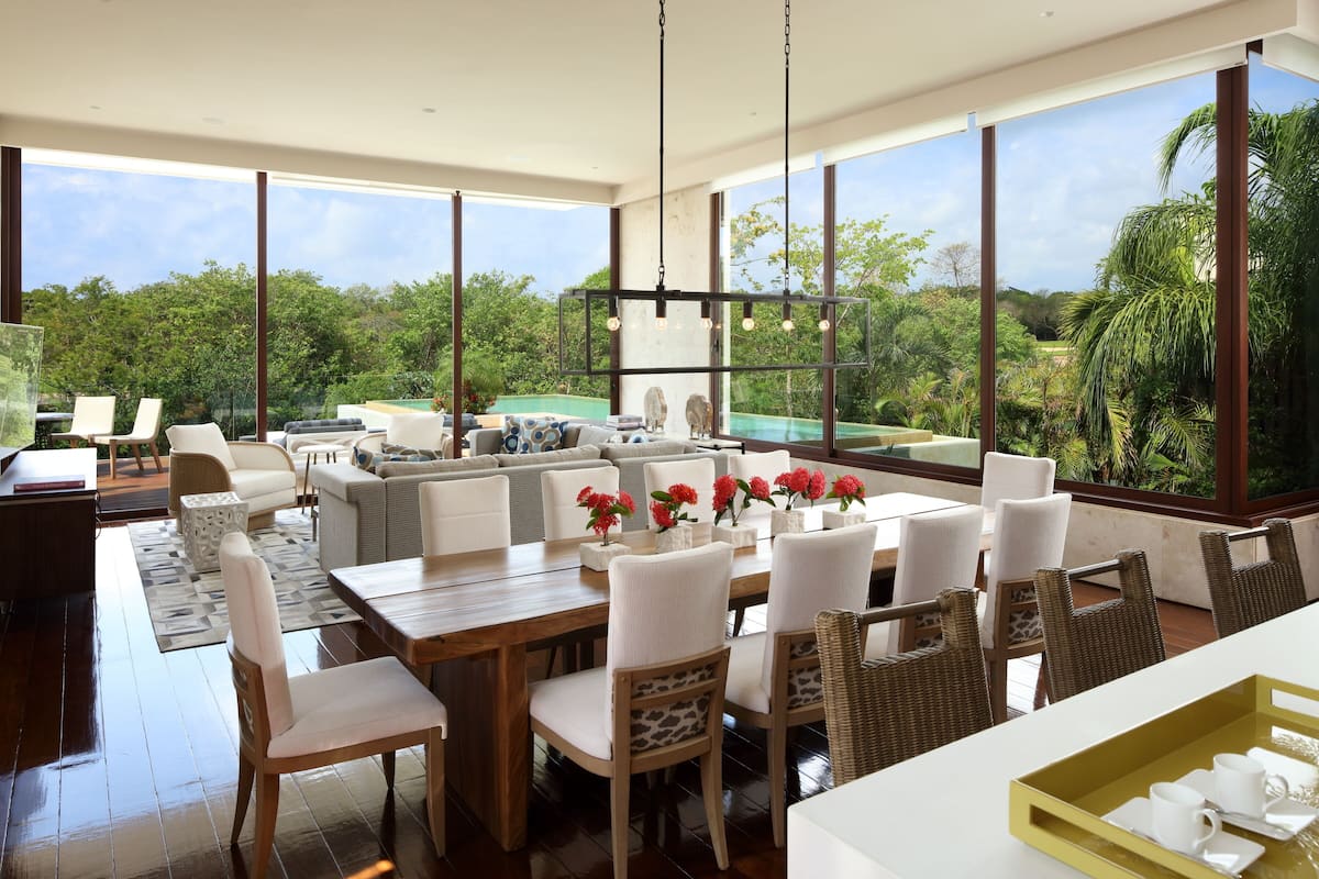 A spacious living area with a large dining table, comfortable seating, and a view of lush greenery through large windows.