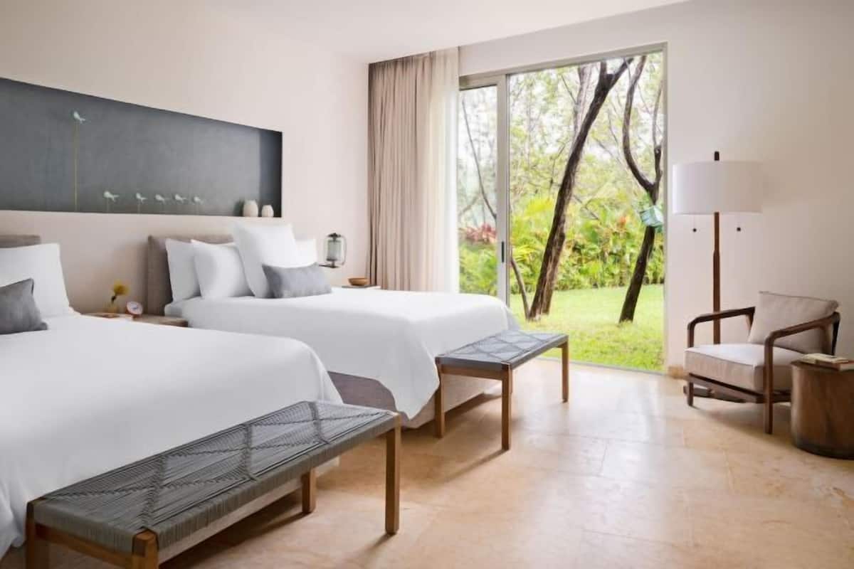 A hotel room with two beds, a bench, a chair, a lamp, and a view of greenery through a sliding door.