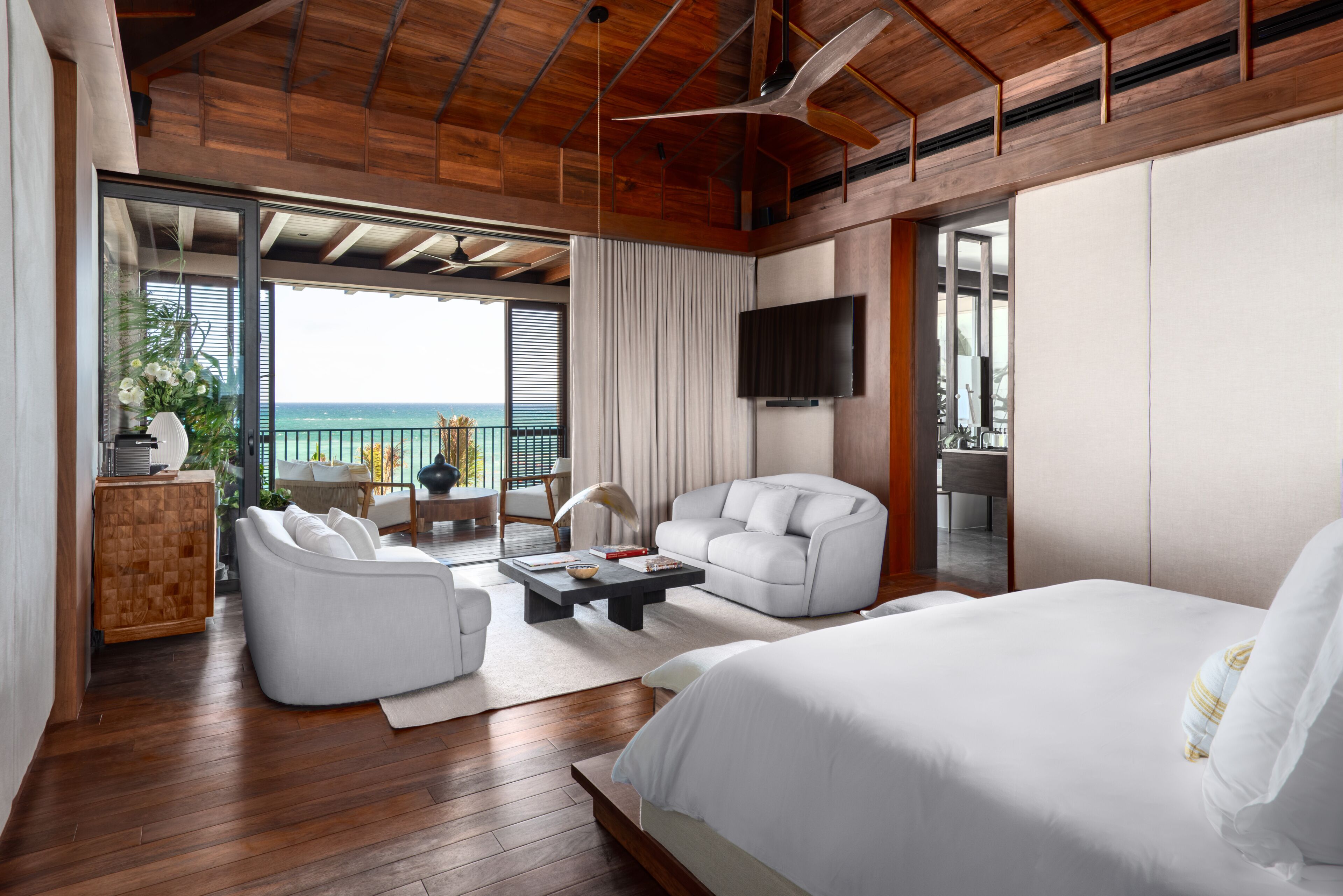 A modern hotel room with a large bed, a sofa, a coffee table, and a TV. There is a balcony with a view of the ocean.