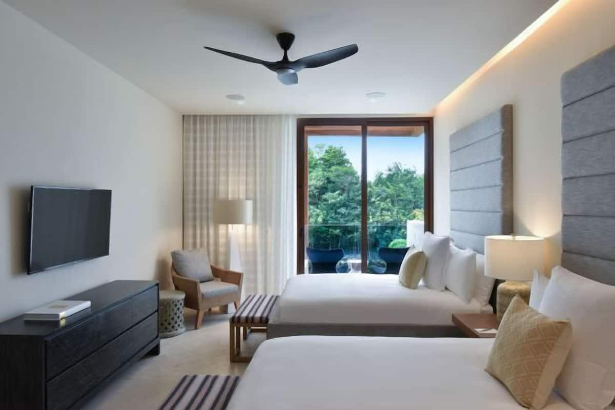 A hotel room with two beds, a TV, a ceiling fan, a window with a view of greenery, and a lamp.