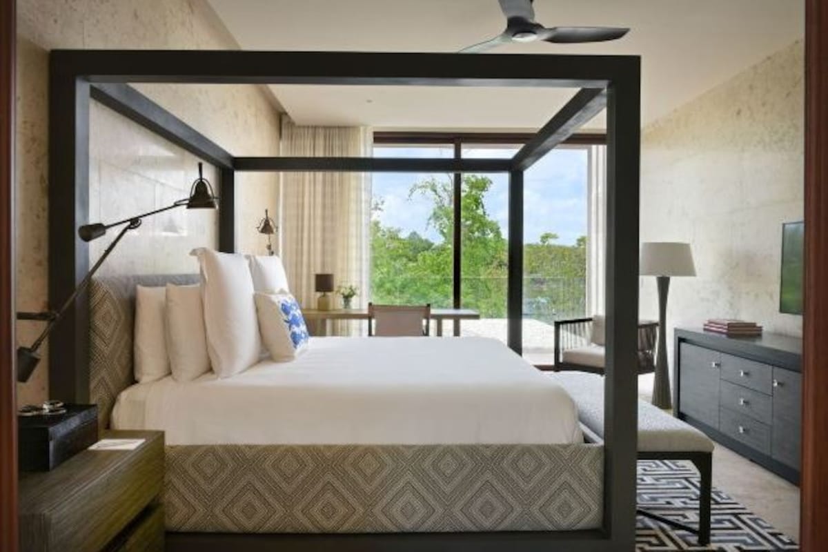 Una habitación de hotel moderna con una cama grande, un escritorio, una silla, una mesita y un balcón con vistas a zonas verdes.