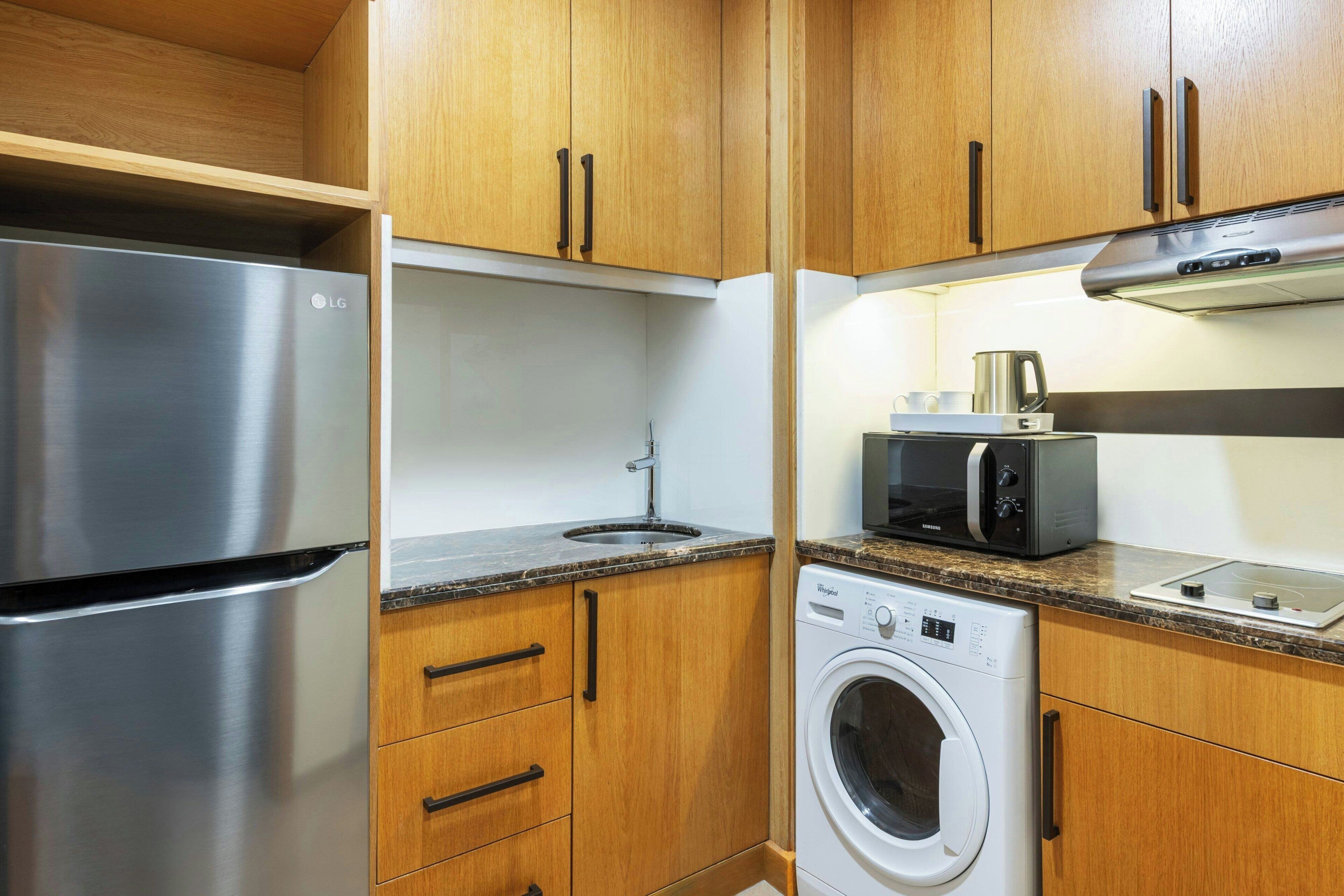 Ett kompakt kök med träskåp, ett kylskåp i rostfritt stål, en mikrovågsugn, en brödrost och en tvättmaskin.