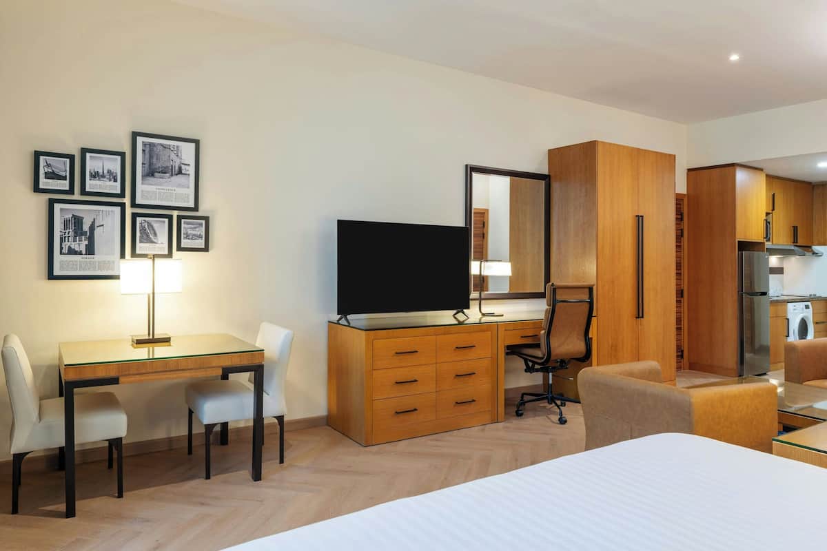 A modern hotel room with a wooden desk, a television, a dining area, and a kitchen area.