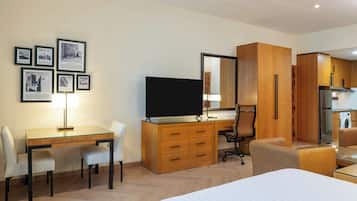 A modern hotel room with a wooden desk, a television, a dining area, and a kitchen area.