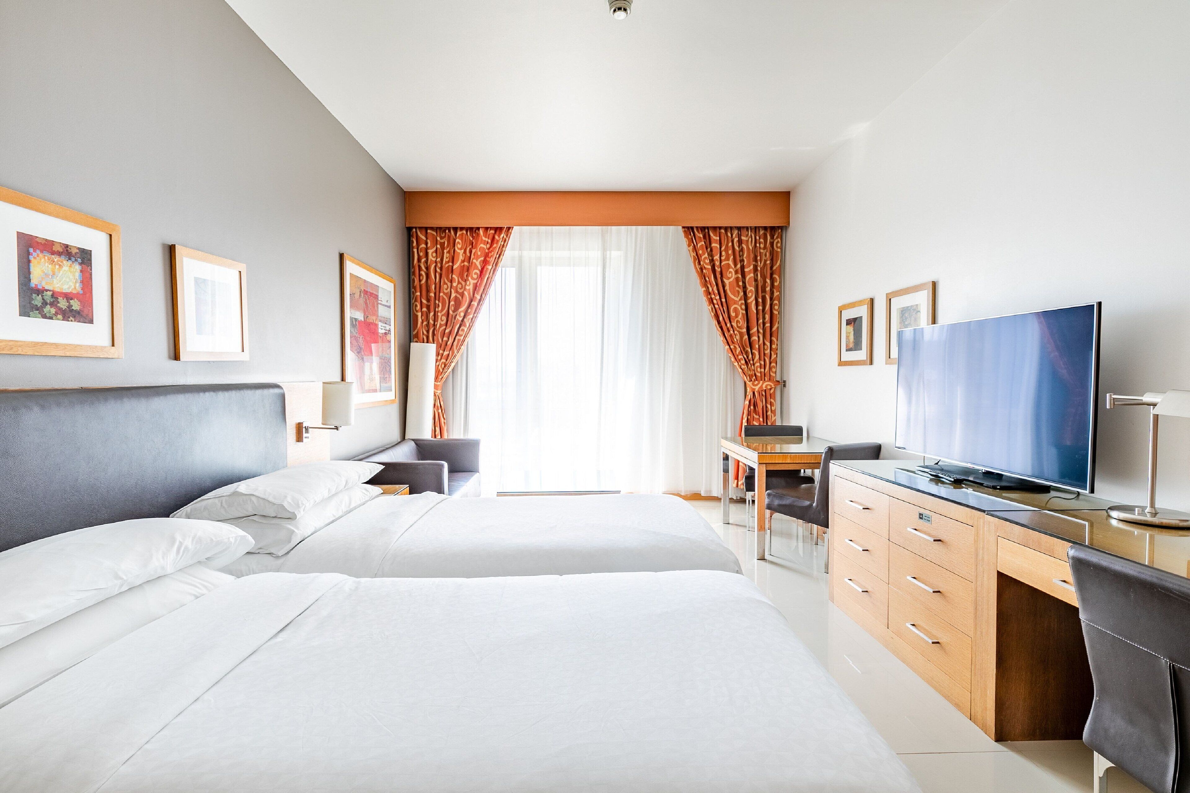 A hotel room with two beds, a desk, a TV, and framed pictures on the wall.