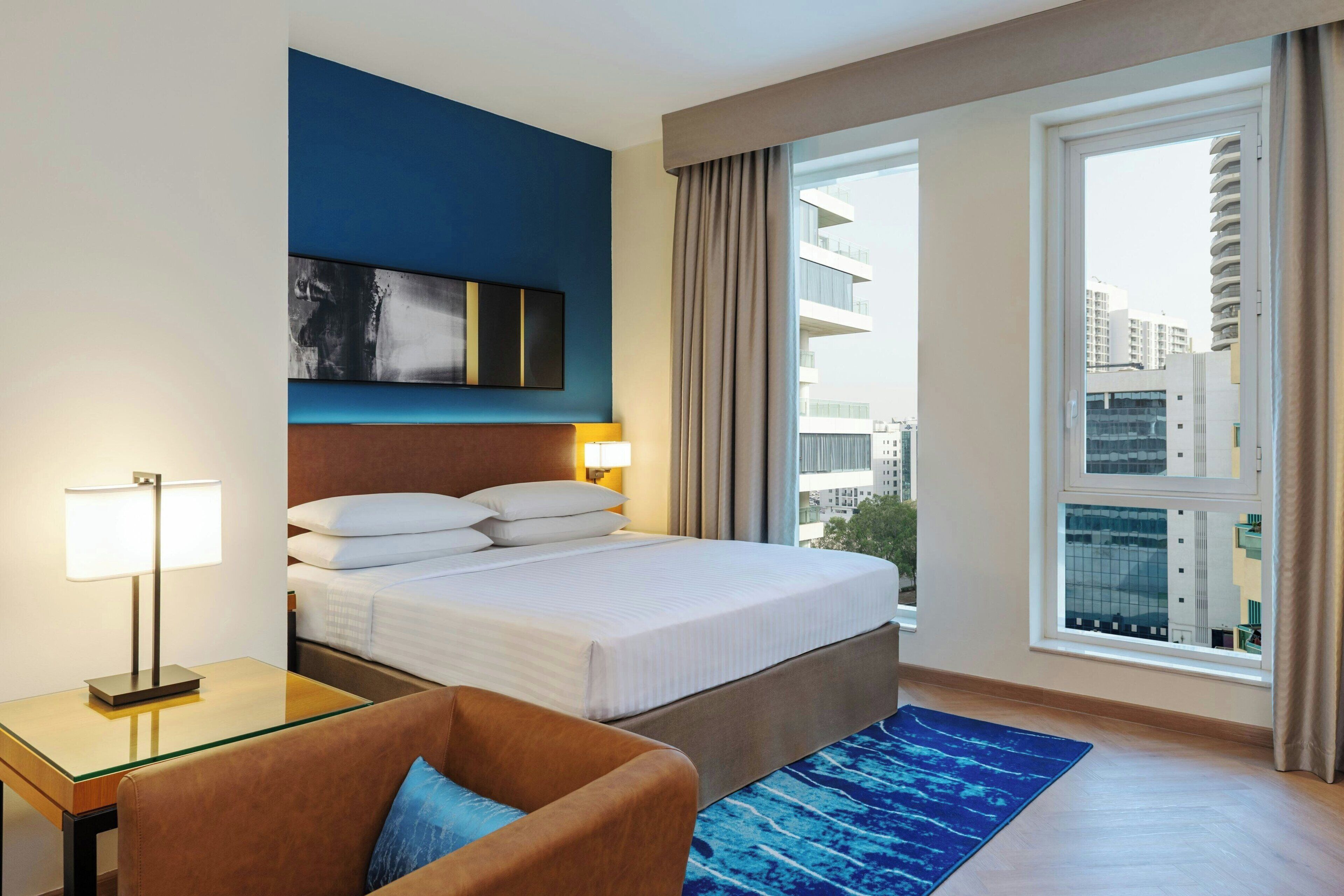 Ein Hotelzimmer mit einem Bett, einem Schreibtisch, einem Stuhl, einem großen Fenster mit Blick auf Gebäude und einem blauen Teppich.