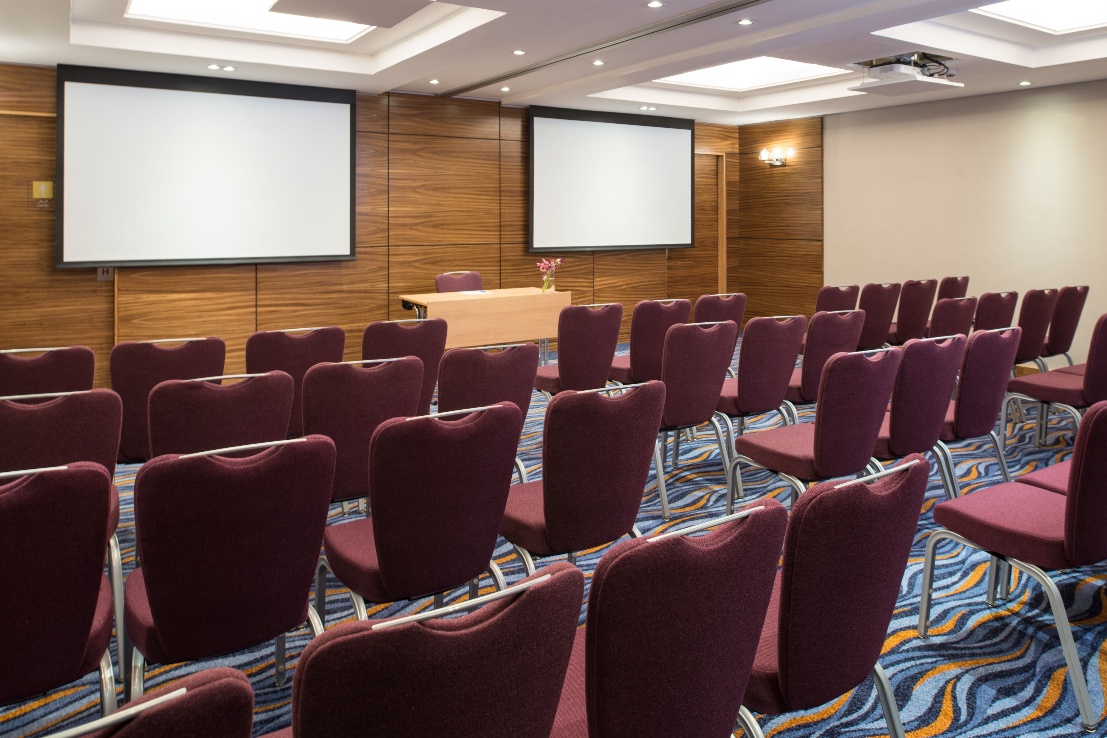 A conference room with rows of purple chairs, a wooden panel wall, a projector screen, and a carpeted floor.
