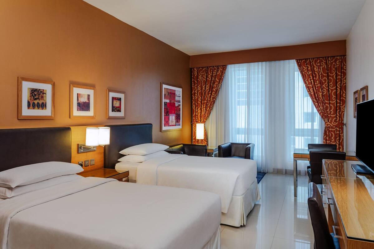 A hotel room with two beds, a desk, a chair, a TV, and framed artwork on the wall.