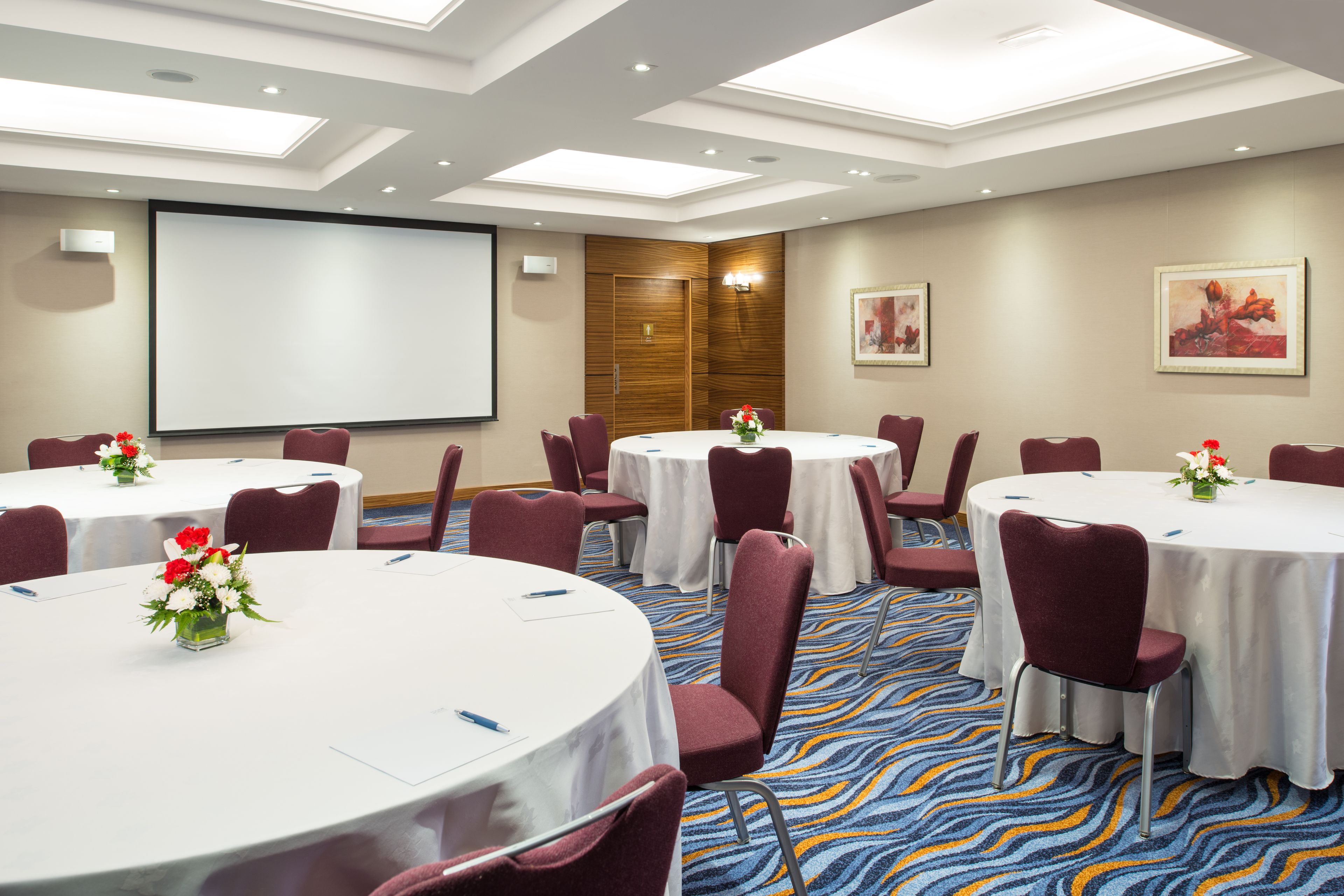 A conference room with multiple round tables, each with a white tablecloth and red chairs. There is a large projection screen on the wall, and the room has a modern ceiling design with skylights.