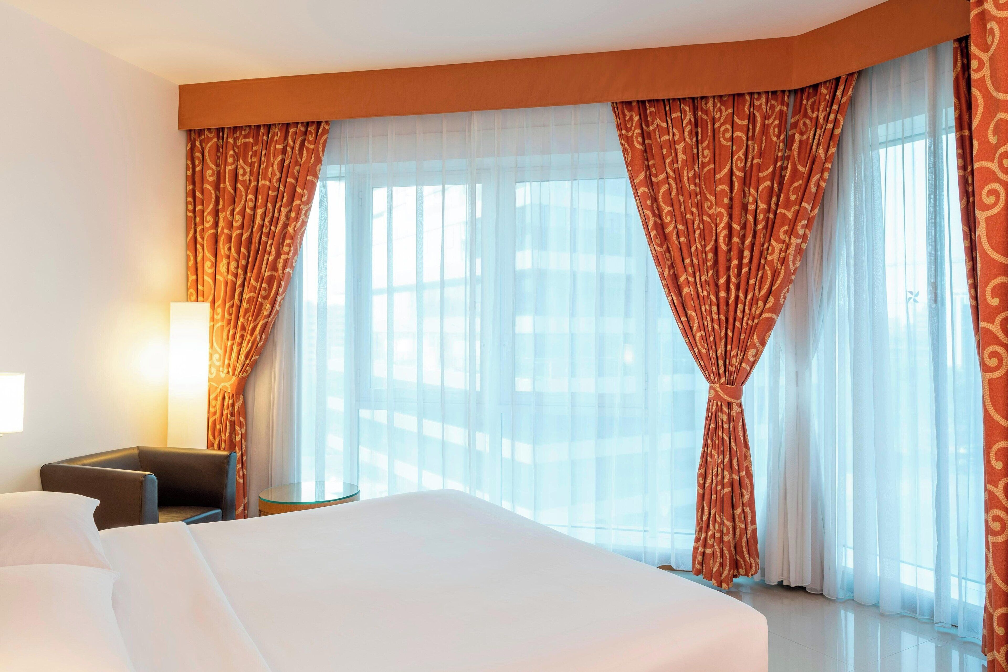 A hotel room with a large bed, a chair, and a small table. There are curtains and a window with a view.