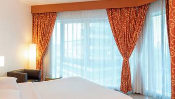A hotel room with a large bed, a chair, and a small table. There are curtains and a window with a view.