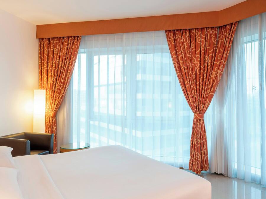 A hotel room with a large bed, a chair, and a small table. There are curtains and a window with a view.