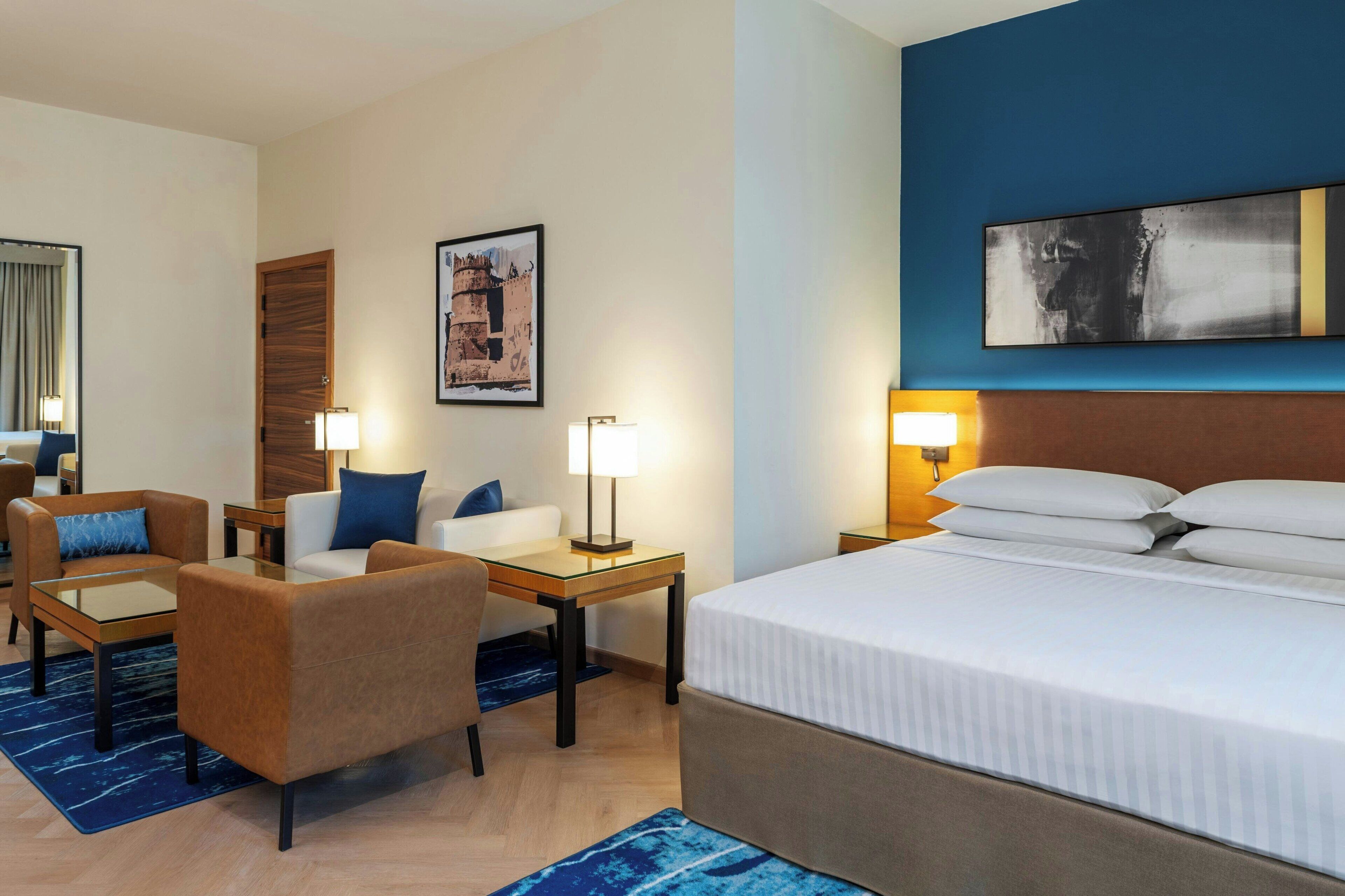 A hotel room with a bed, two armchairs, a small table, a desk, and a nightstand with lamps.