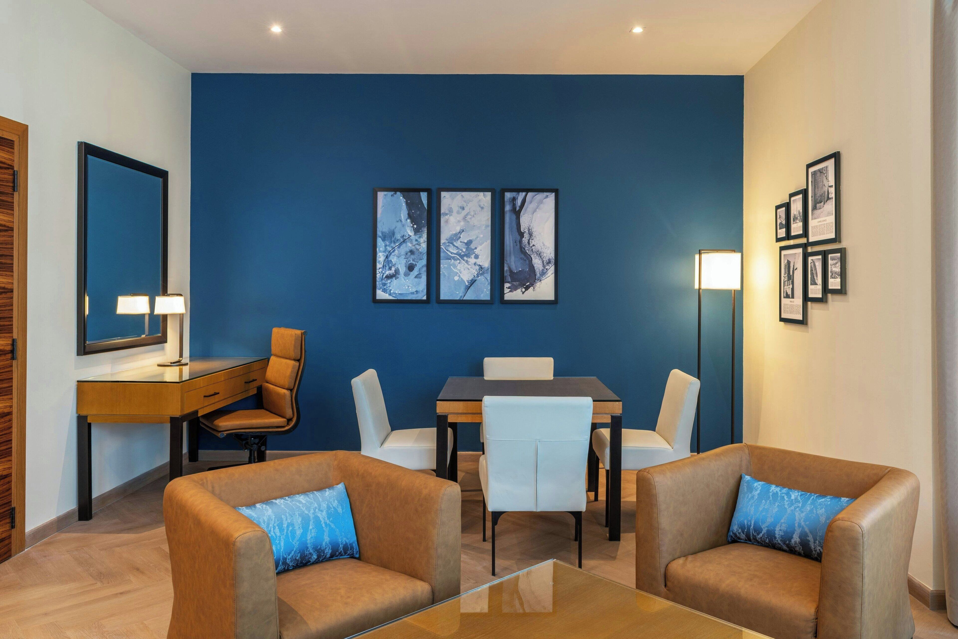 A modern hotel room with a blue accent wall, a dining area with a table and chairs, and a seating area with two sofas.