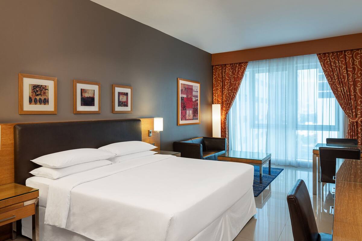 A hotel room with a large bed, a desk, a chair, a small table, and a window with curtains. There are framed pictures on the wall.