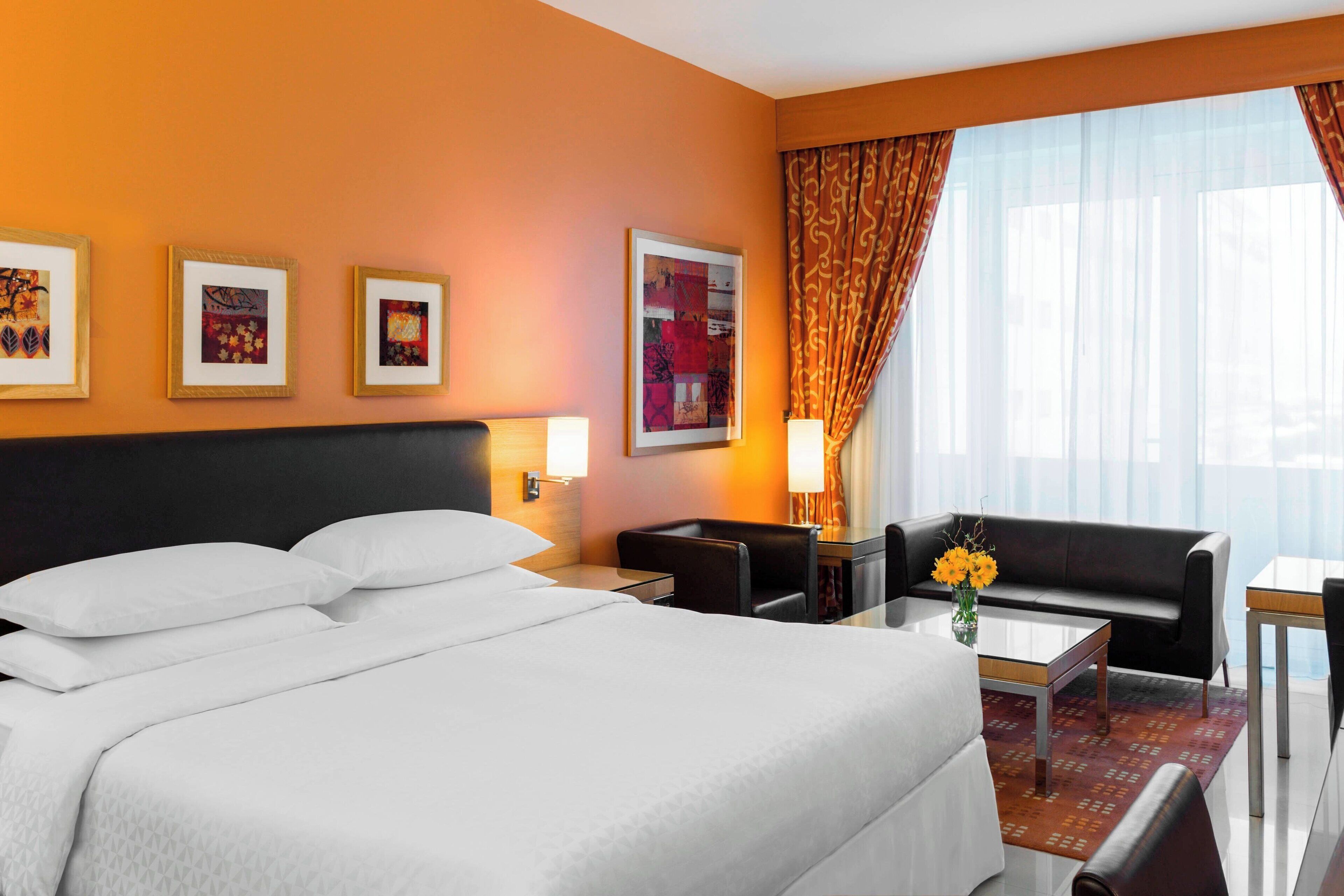 A hotel room with a large bed, a sofa, a small table, and a desk. There are framed pictures on the wall and a window with curtains.