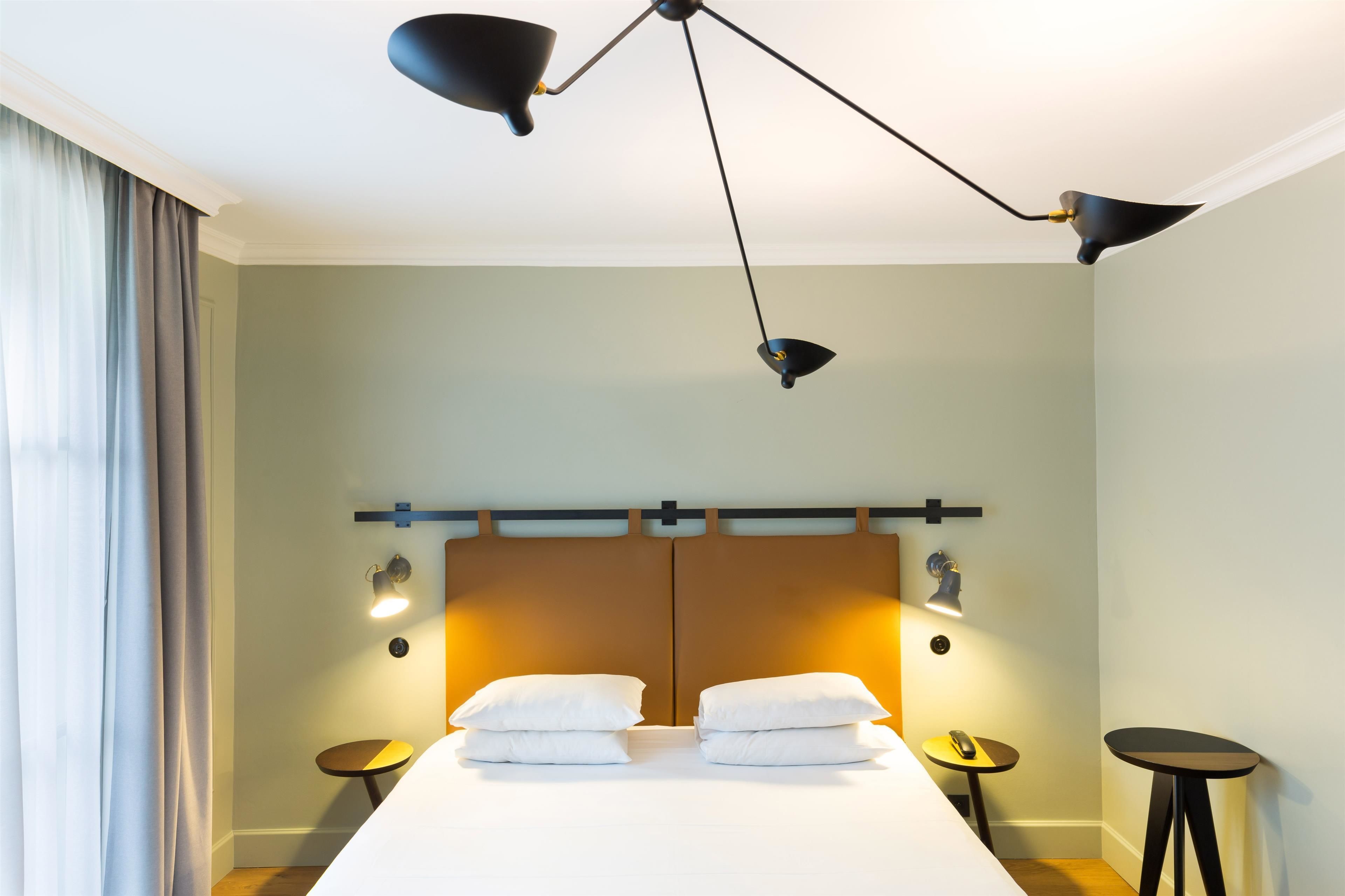 Una habitación de hotel con una cama grande, dos mesitas de noche, una lámpara de techo moderna y una ventana con cortinas.