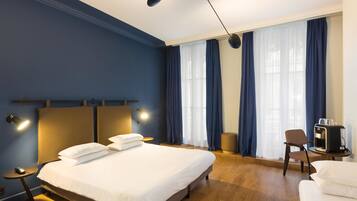 Ein Hotelzimmer mit einem großen Bett, einem Schreibtisch mit Stuhl, einer Kaffeemaschine und einem Fenster mit Vorhängen.