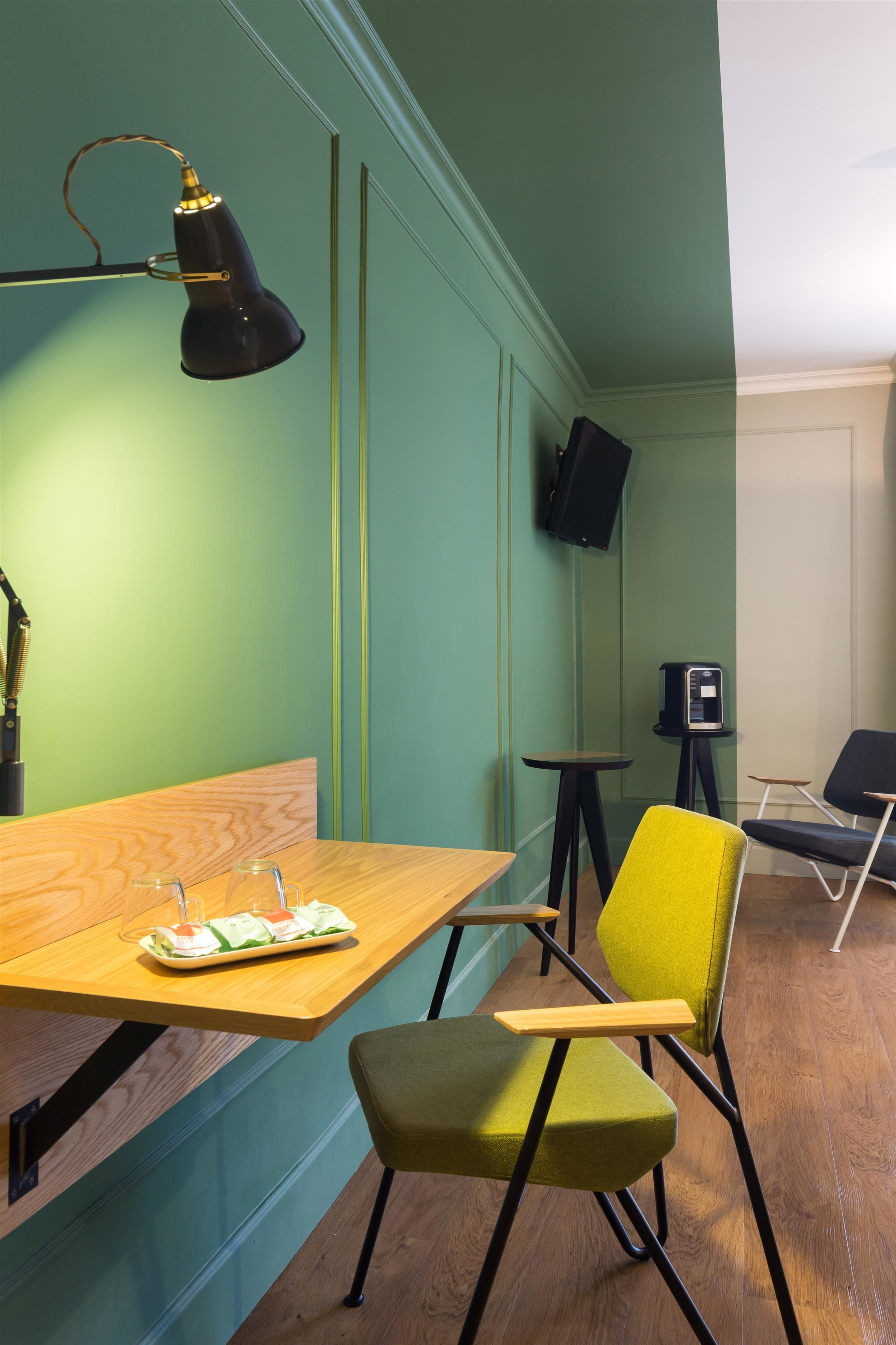 A room with a wooden table, a green chair, a black desk lamp, a speaker, and a television mounted on the wall.