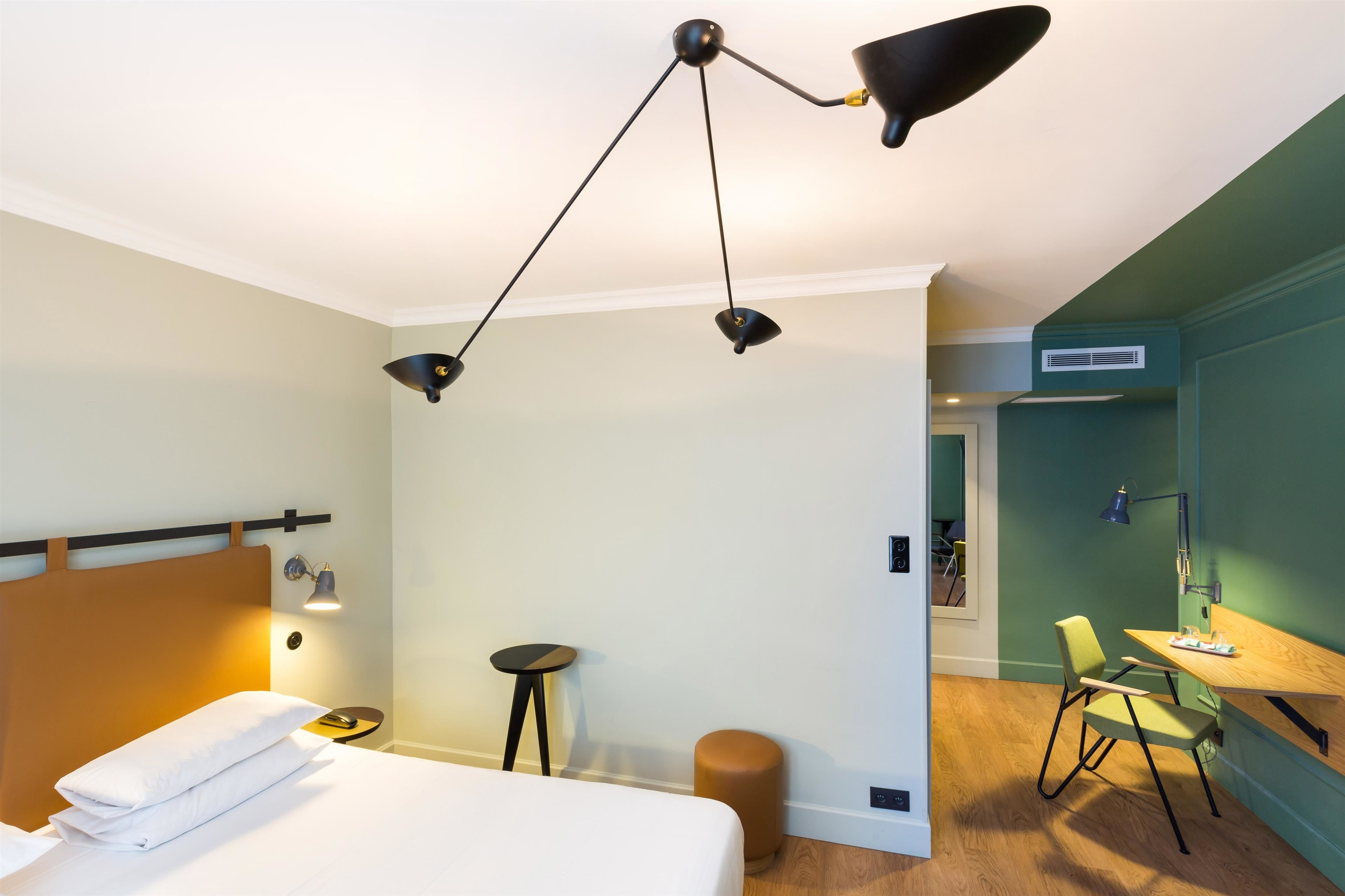 Ett modernt hotellrum med en säng, ett sängbord, en lampa och en liten pall.