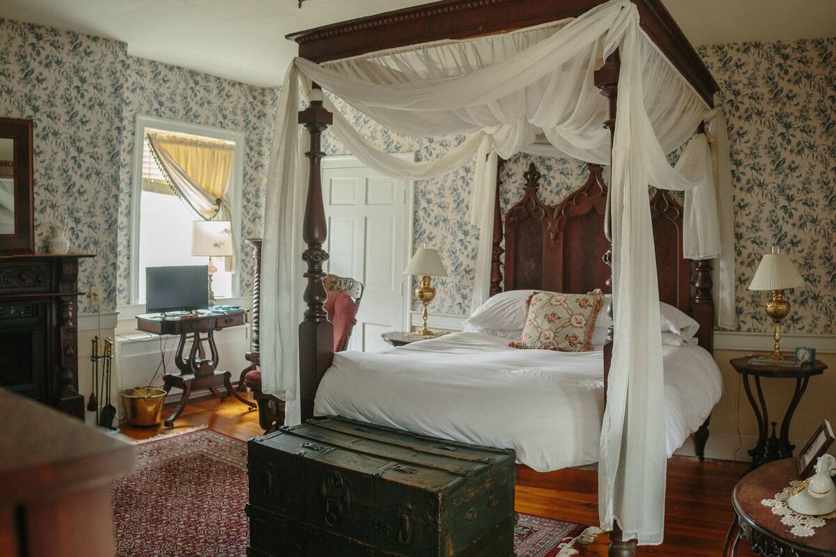 A four-poster canopy bed with white linens, a vintage-style lamp, and a wooden chest in a room with floral wallpaper and a red patterned rug.