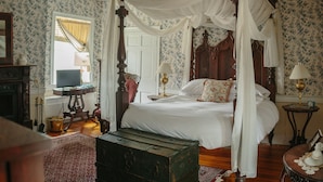 A four-poster canopy bed with white linens, a vintage-style lamp, and a wooden chest in a room with floral wallpaper and a red patterned rug.